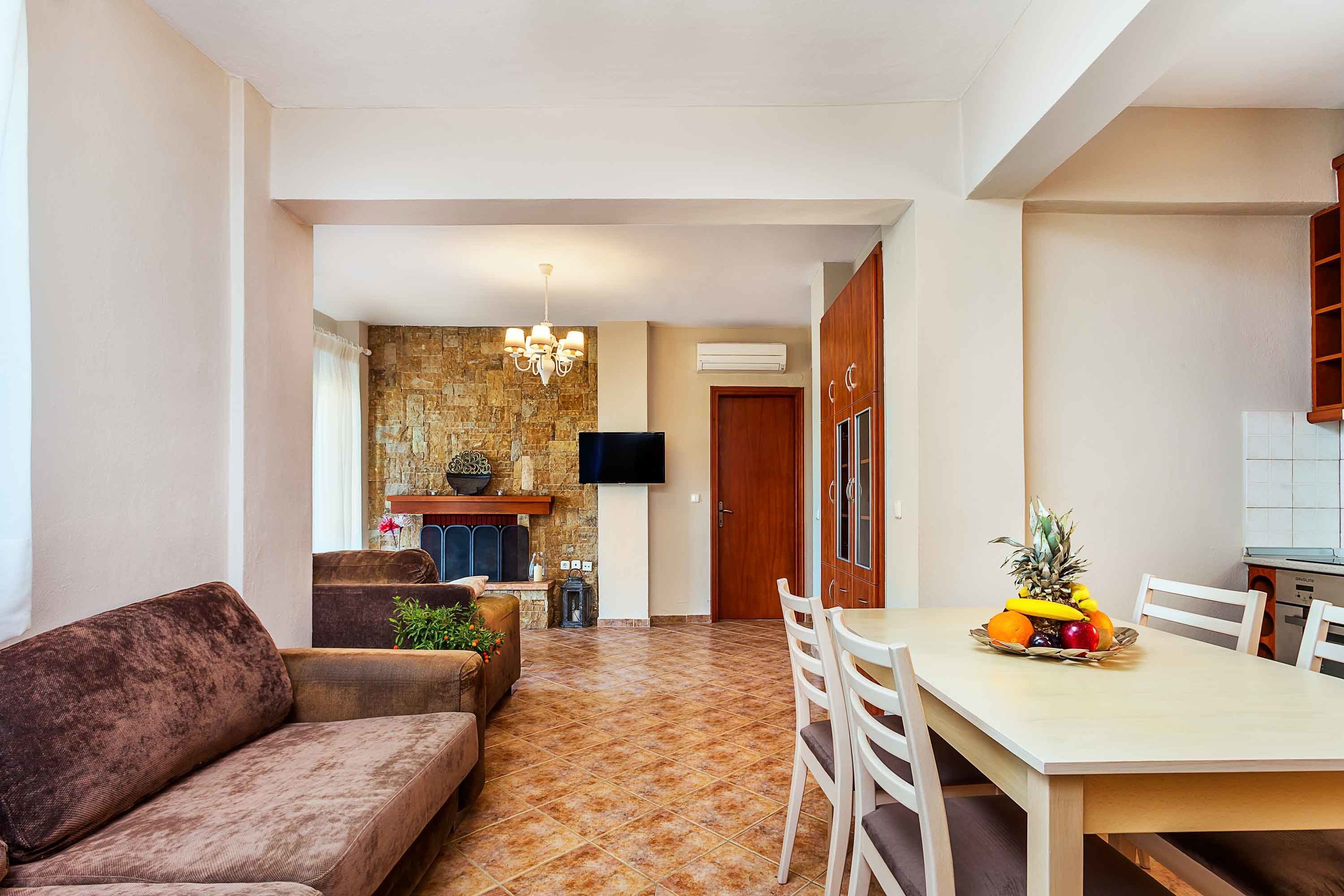 Un salon avec un canapé marron, une table à manger avec des chaises et un mur en pierre sur lequel est accrochée une télévision.