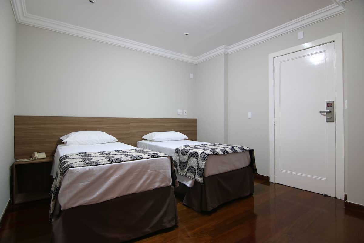 Une chambre d’hôtel avec deux lits, une tête de lit en bois, une porte blanche et une petite table de chevet avec un téléphone.