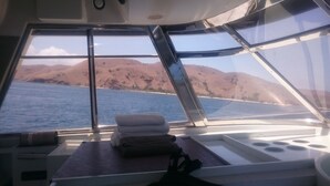 A boat interior with a view of the sea and mountains. There are towels stacked on a table, and a bag is on the floor. - Laislabonita (Labuan Bajo)