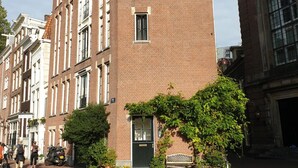 Ein mehrgeschossiges Backsteingebäude mit einem zentralen Eingang, großen Fenstern und einer kleinen Gartenanlage mit Kübelpflanzen.
