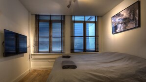 Ein Schlafzimmer mit einem Bett, zwei Fenstern mit Jalousien, einem an der Wand montierten Fernseher und einem gerahmten Bild.