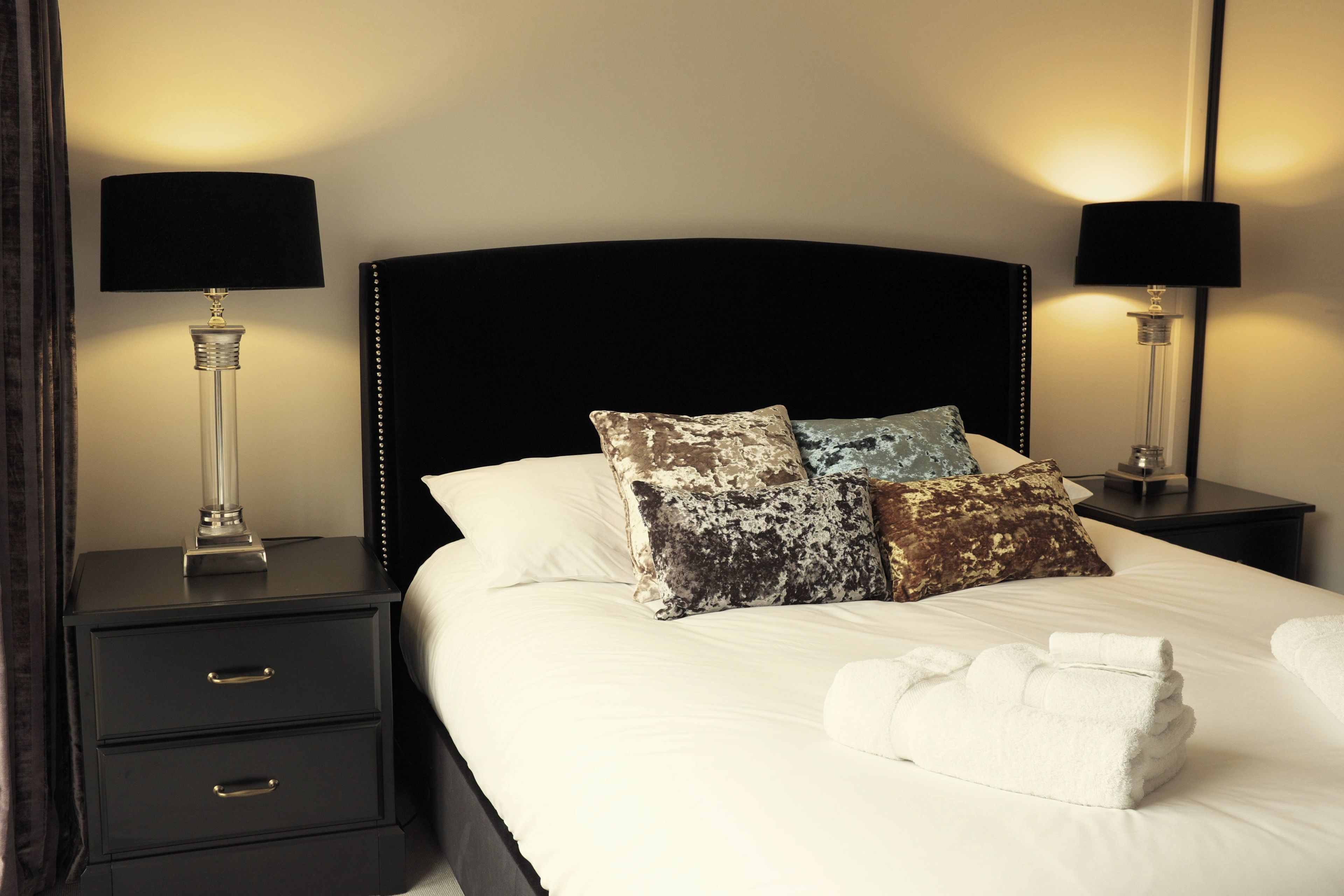 A neatly made bed with a black headboard, two bedside tables with lamps, and a stack of white towels.