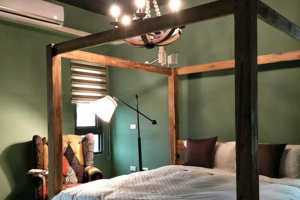 A four-poster bed with a wooden frame and white bedding, a chandelier, and a window with blinds.