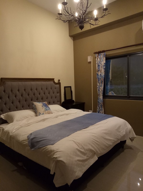 Un dormitorio con una cama grande, cabecera capitonada, mesita de noche, espejo, ventana con cortinas y una araña.