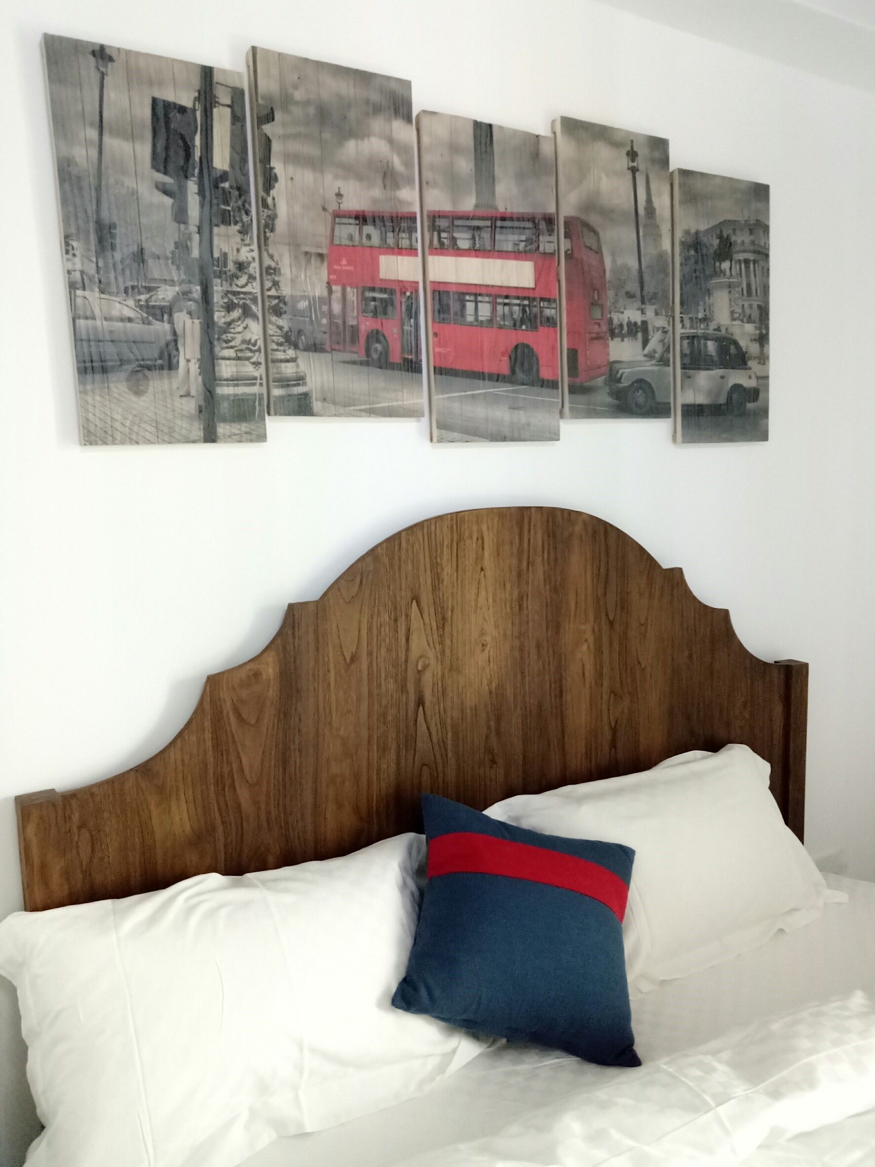 A bed with white linens and a blue and red pillow, a wooden headboard, and four framed pictures of a red double-decker bus on the wall above.