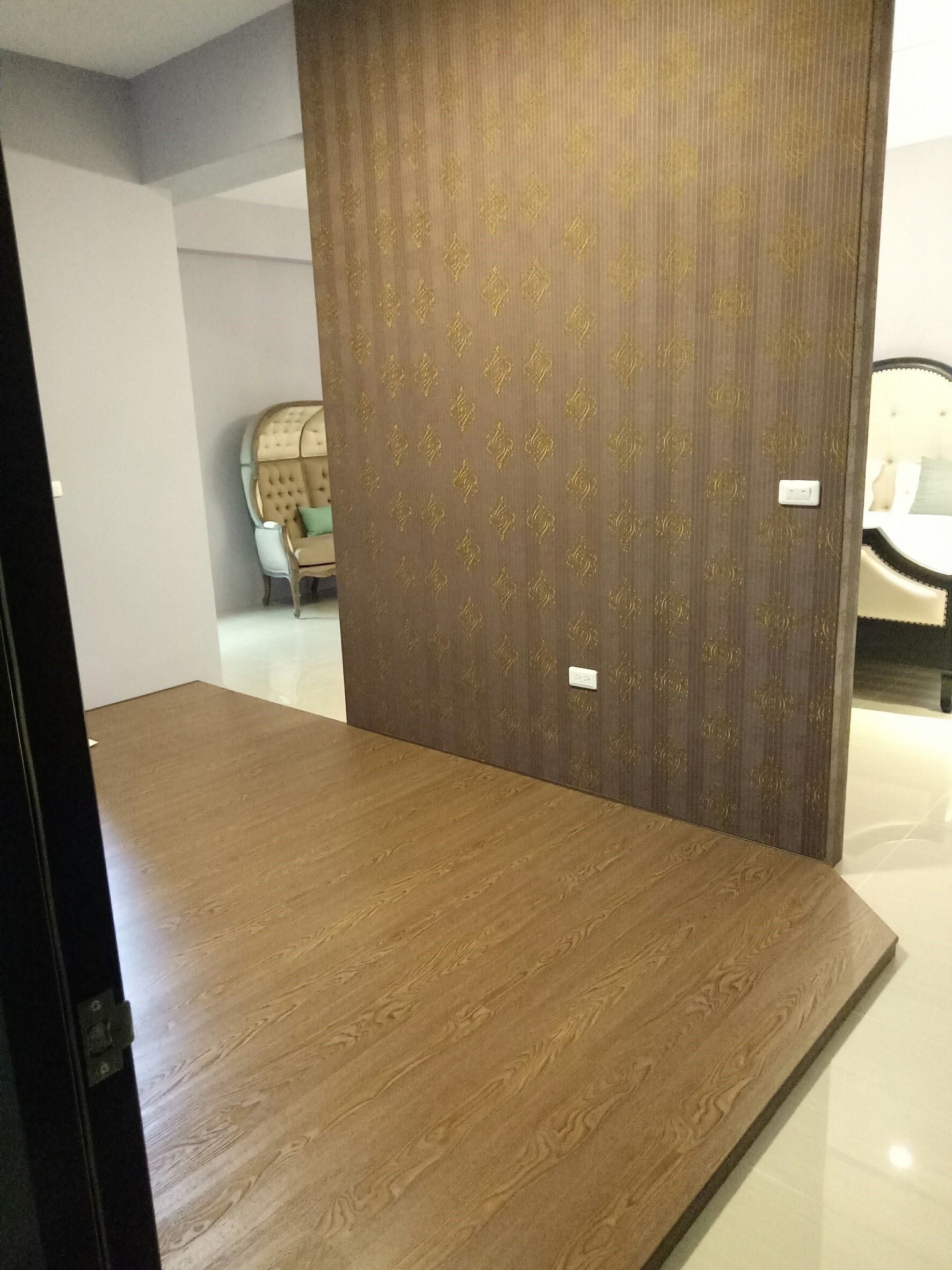 A room with a wooden floor, a dark patterned partition, and a bed with a white headboard.