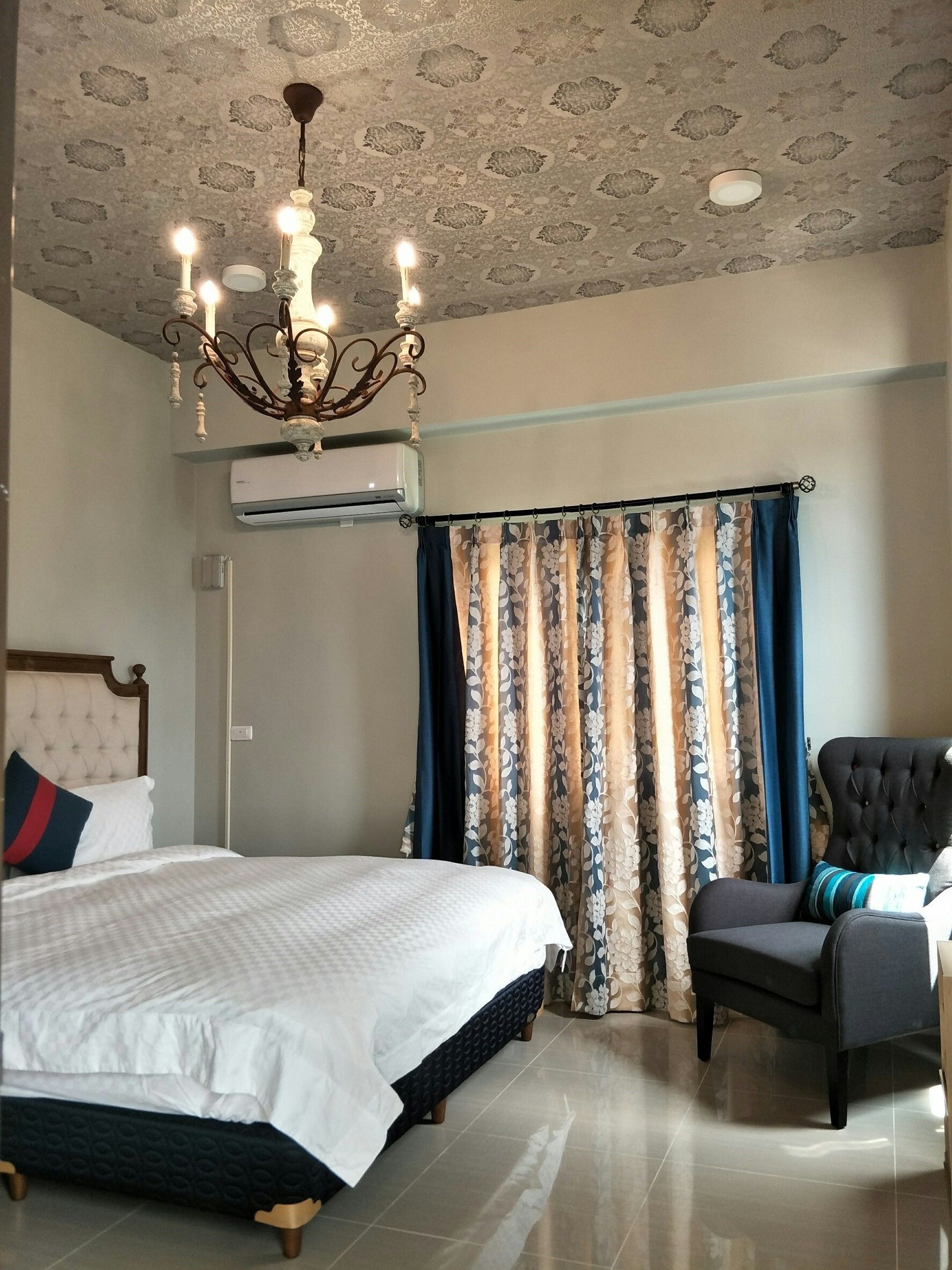 A hotel room with a bed, a chair, a chandelier, and a window with curtains.