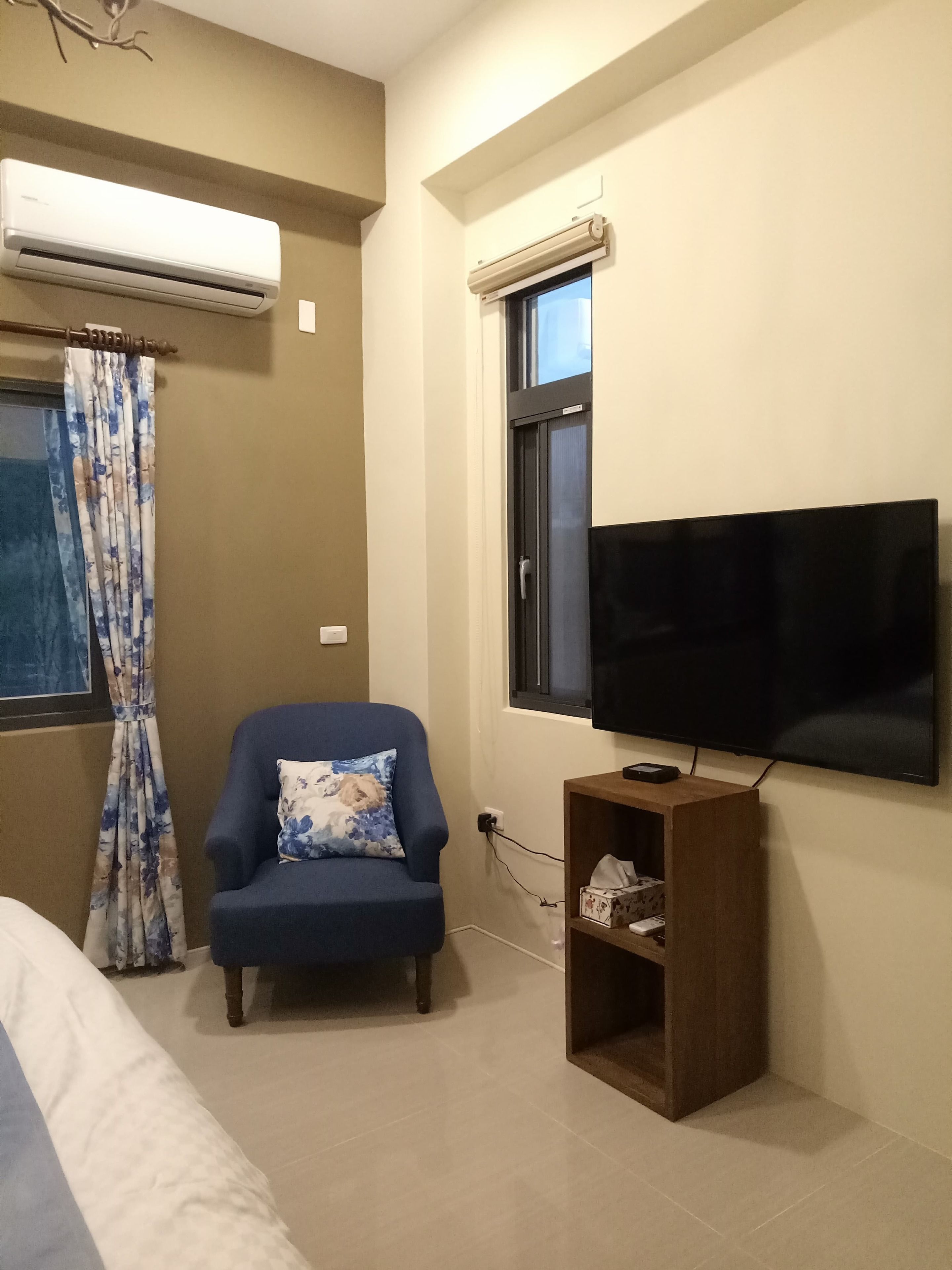 A room with a blue armchair, a flat-screen TV mounted on the wall, an air conditioner, and a window with a curtain.