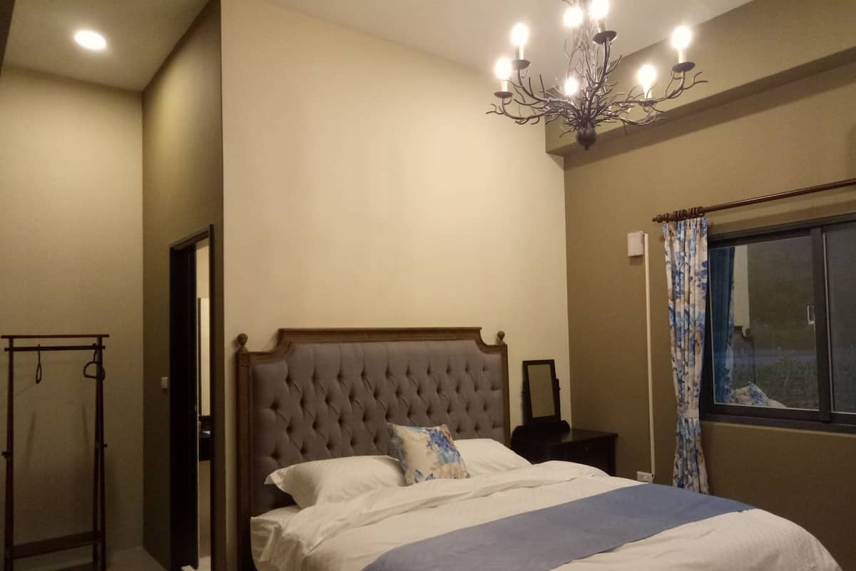 A bedroom with a large bed, a chandelier, a window with curtains, and a bedside table.