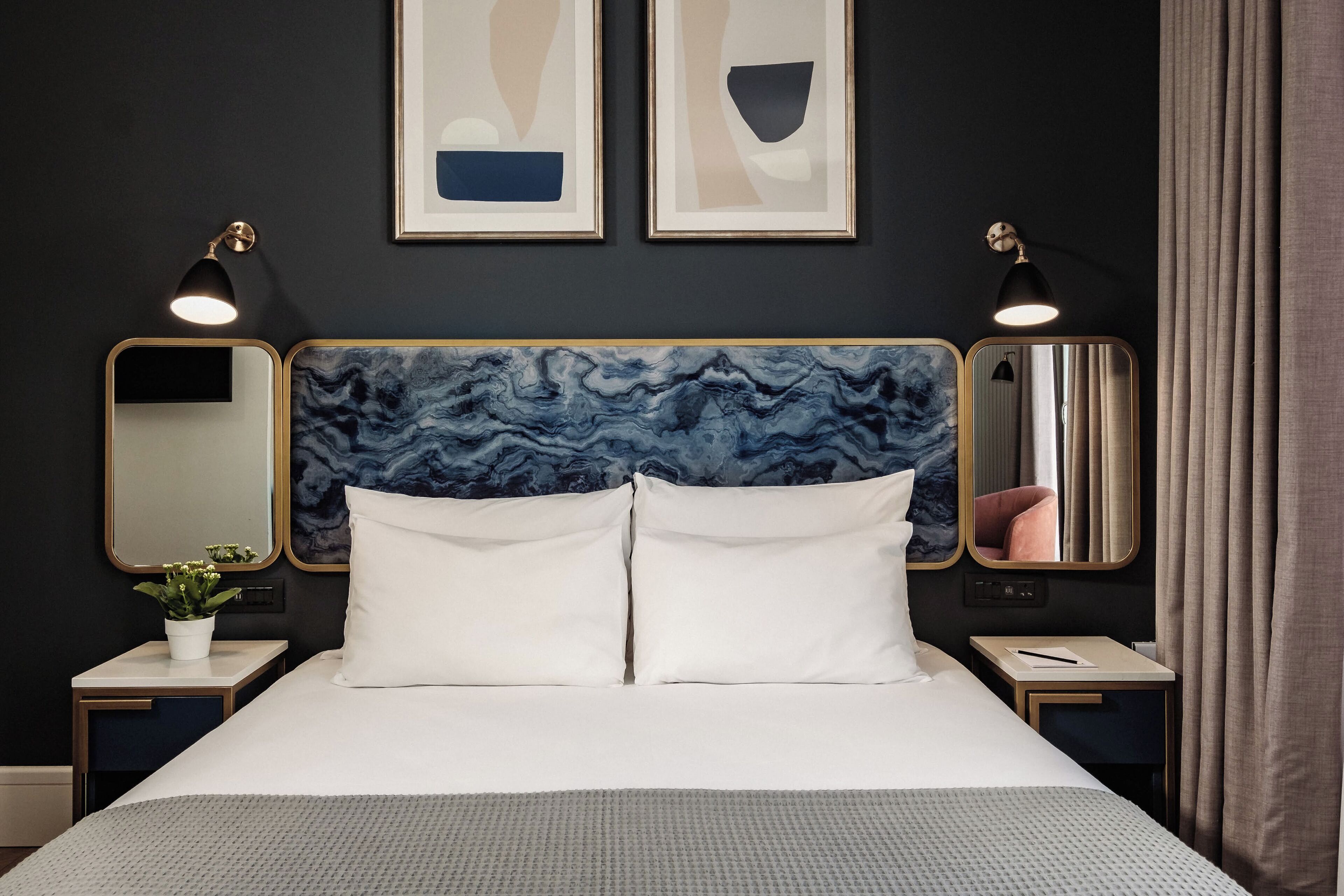 A hotel room with a large bed, two bedside tables, a nightstand with a plant, two wall-mounted mirrors, and abstract artwork above the bed.