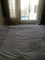A bedroom with a large bed, a window with a view of a swimming pool, and sheer curtains.