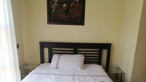 Un lit bien fait, avec du linge de lit blanc et une housse de lit grise à motifs, une seule serviette turquoise et un tableau au-dessus du lit.