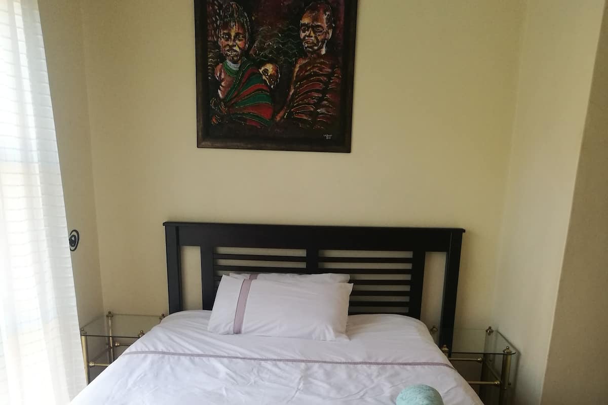 Una cama bien tendida con sábanas blancas y una falda de cama gris estampada, una toalla azul aguamarina y un cuadro sobre la cama.