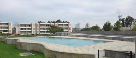 Una piscina rodeada por un solárium de hormigón, con un césped en primer plano y edificios de varios pisos al fondo.