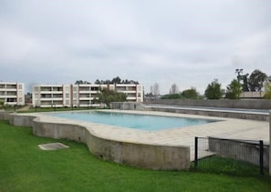 A swimming pool surrounded by a concrete deck, with a grassy area in the foreground and multi-story buildings in the background.