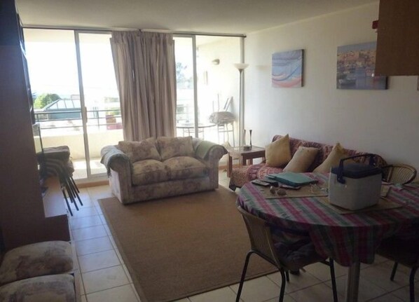 A living room with a sofa, a dining table with chairs, a small table with a lamp, and a view through a large window.