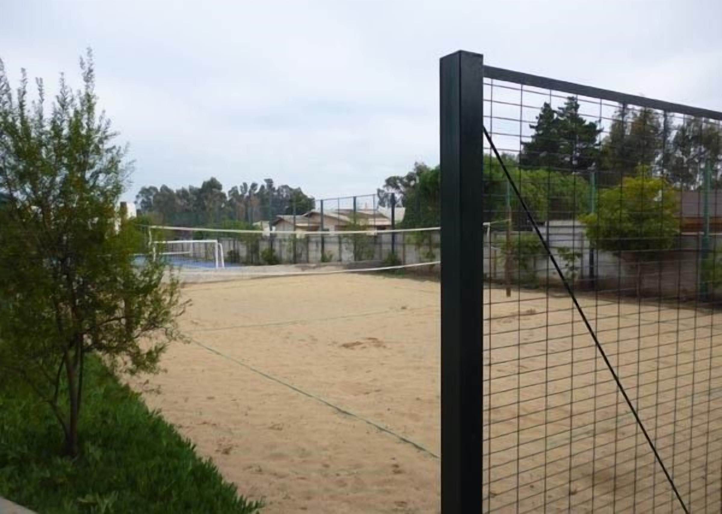 En udendørs volleyballbane med sand, net og hegn, omgivet af træer.