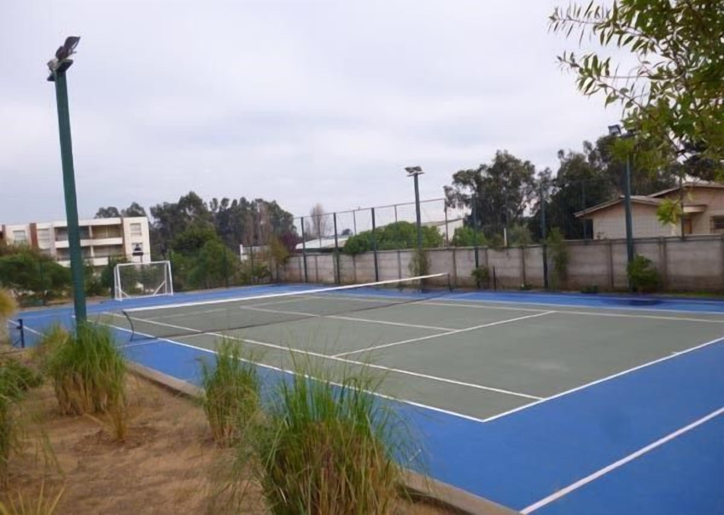 Cancha de tenis exterior con red, rodeada por una cerca y vegetación.