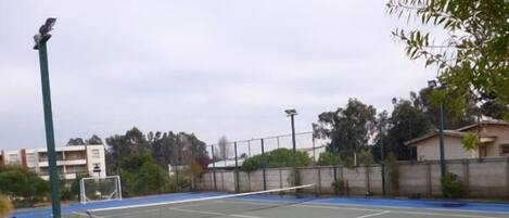 An outdoor tennis court with a net, surrounded by a fence and vegetation.