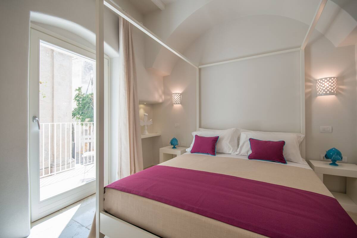 A bedroom with a large bed, two bedside tables, a nightstand, a window with curtains, and a balcony.