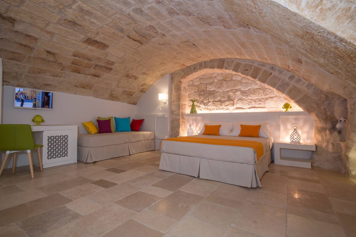 A cozy room with a stone ceiling, a bed with orange bedding, a green chair, and a small table with a lamp.