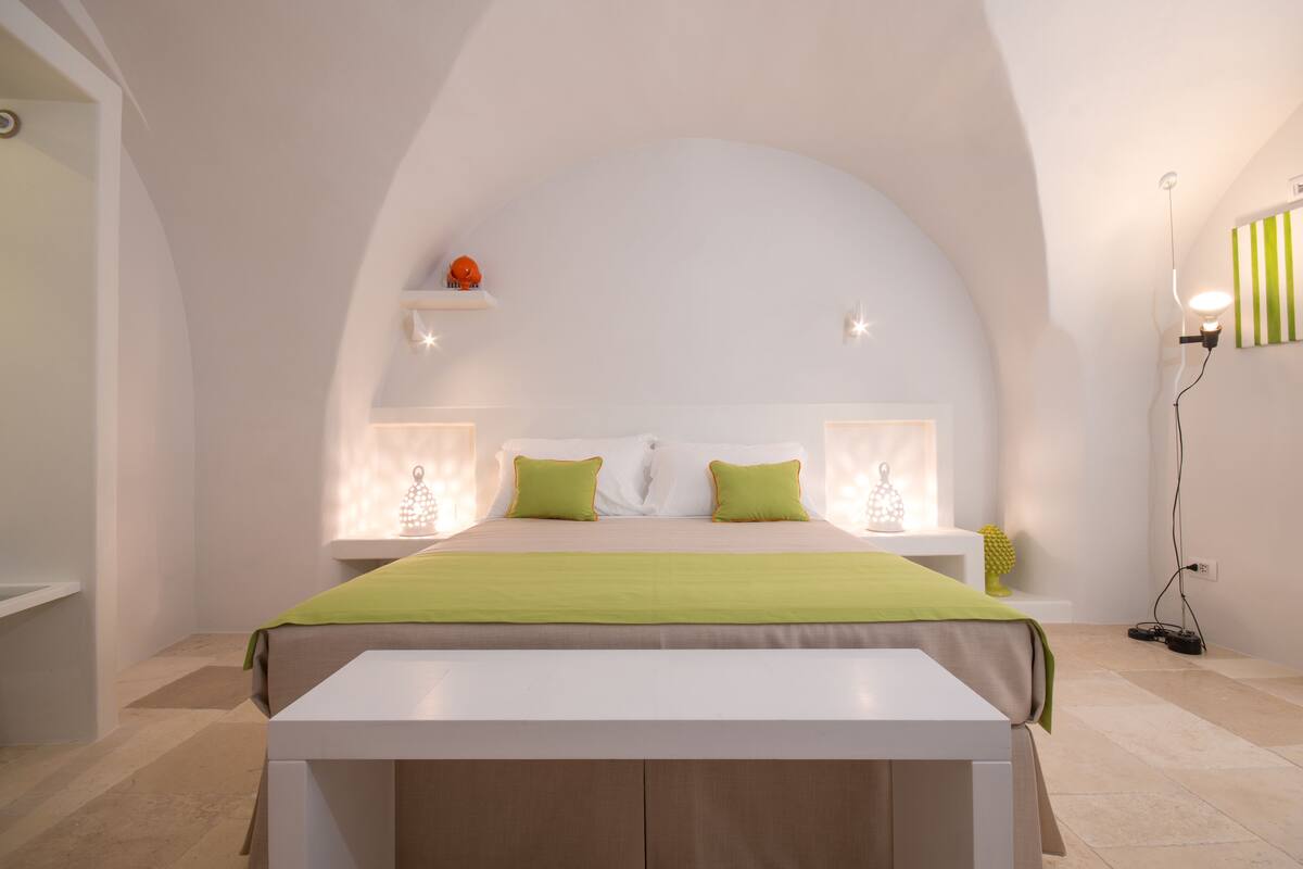 A modern bedroom with a curved ceiling, a bed with green and white bedding, bedside tables with lamps, and a small shelf with a decorative item.