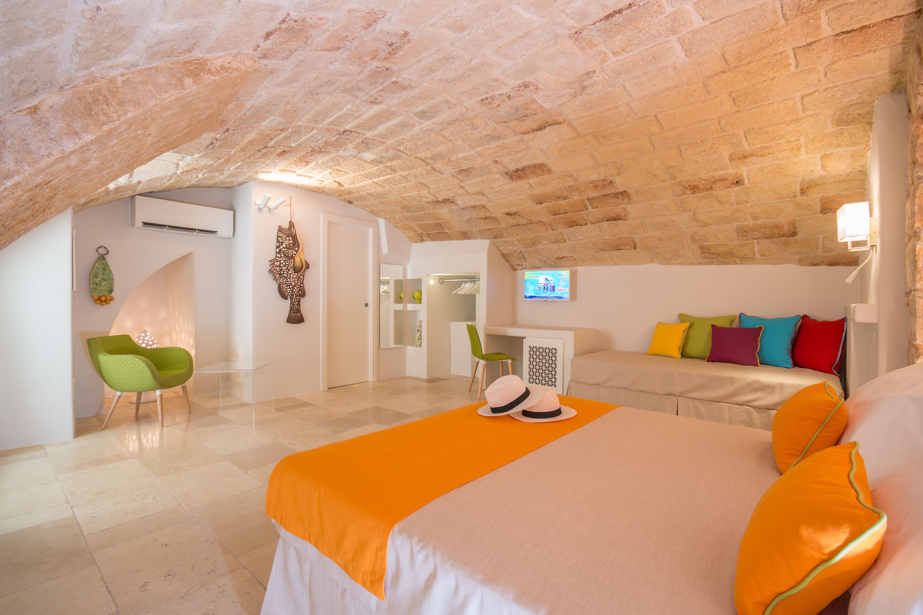 An attic bedroom with a high, exposed brick ceiling, a bed with orange bedding, a green chair, and a small desk with a chair.
