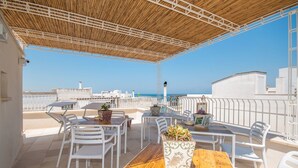 A covered outdoor patio with a wooden table and white chairs, overlooking a clear blue sky and a distant coastline.