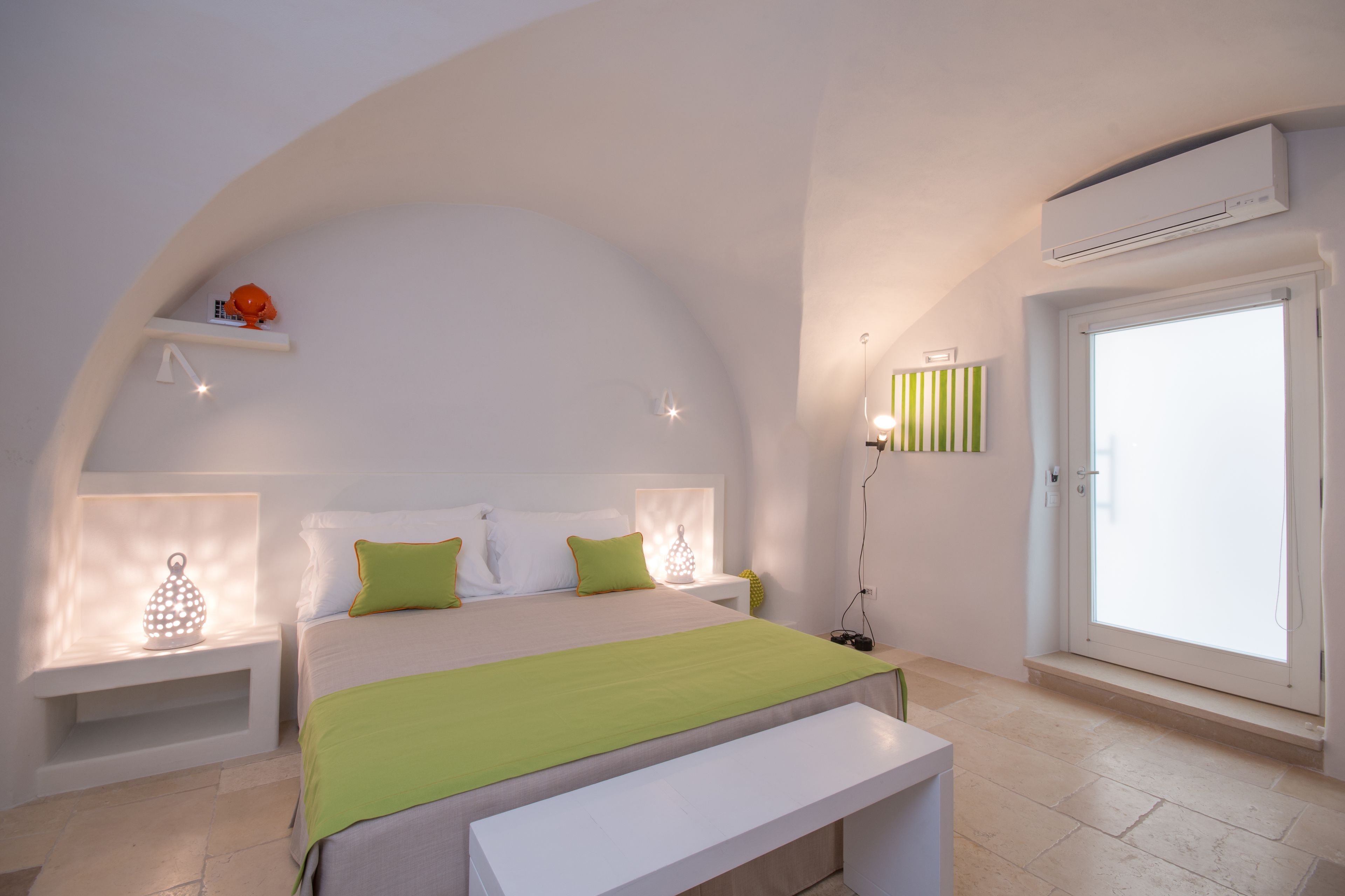 A bedroom with a bed, bedside tables, a bench, an air conditioning unit, and a door.