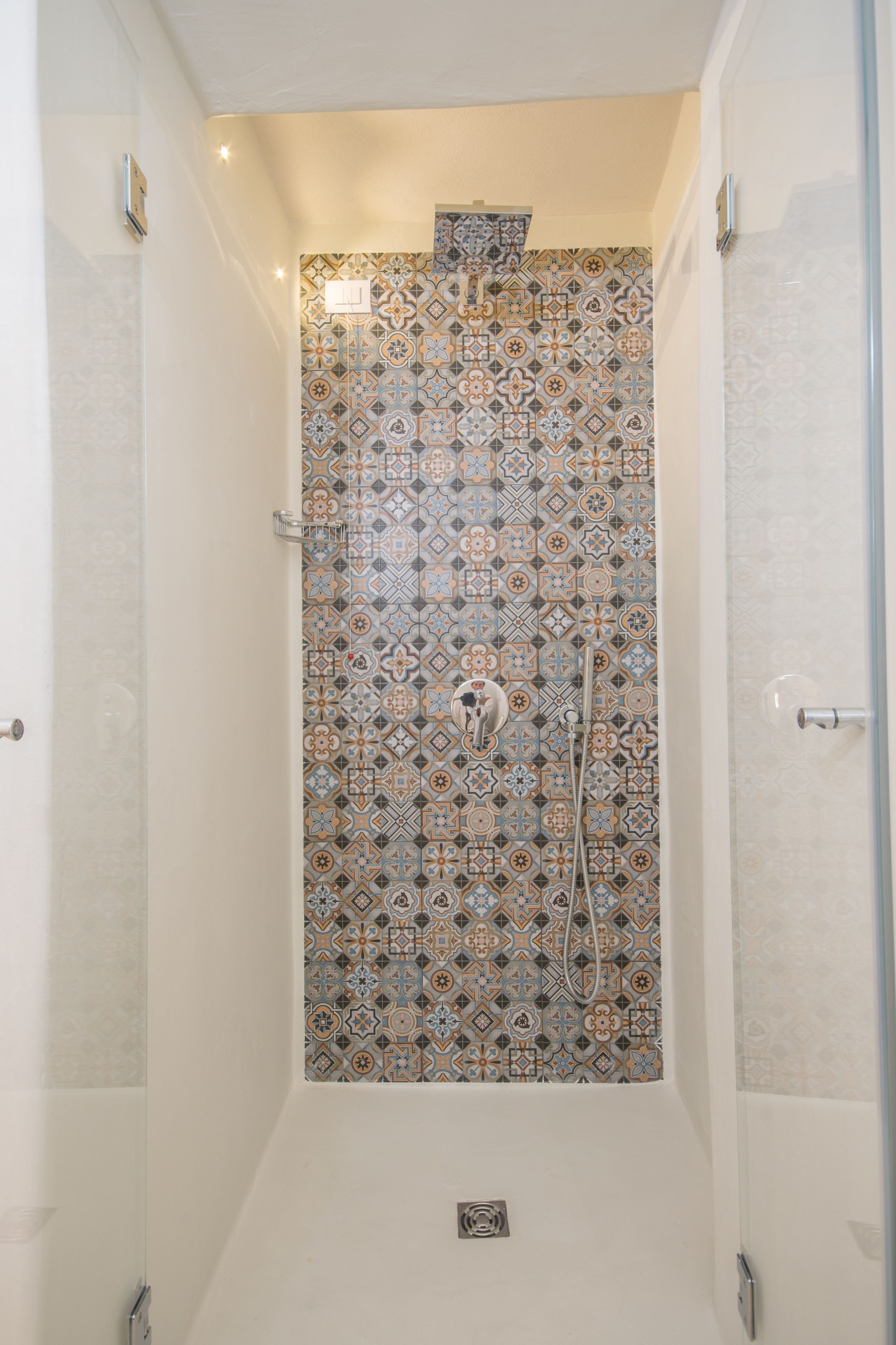 A shower with a patterned tile wall, a visible showerhead, and a small vent on the floor.