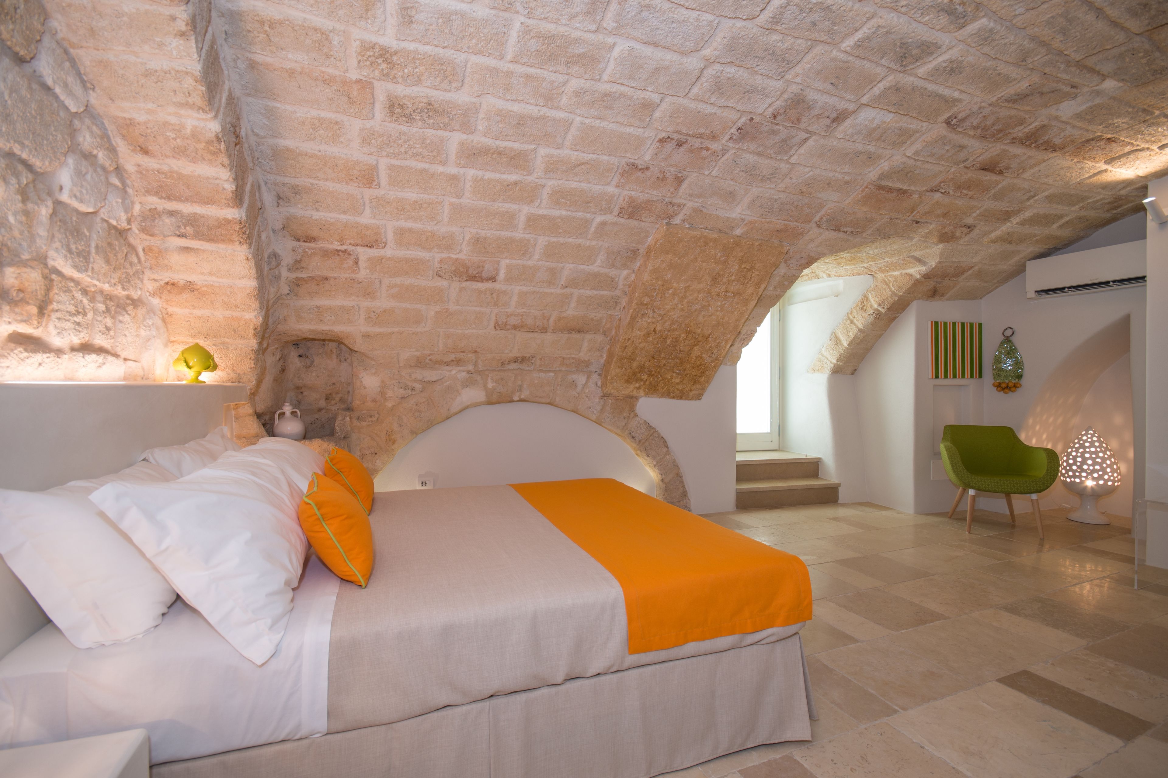 A bedroom with a stone ceiling, a bed with white and orange bedding, a green chair, and a lamp.
