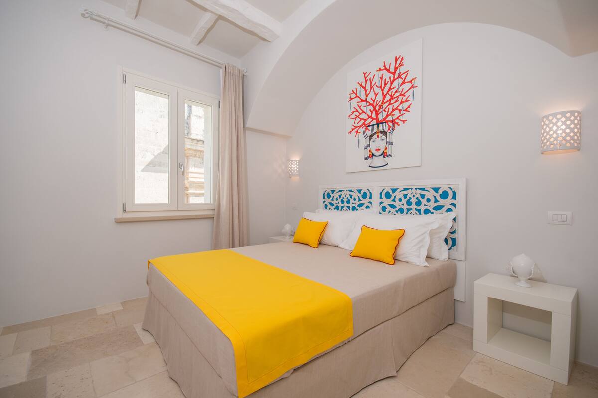A bedroom with a bed, a nightstand, a window, and a wall with a decorative piece.