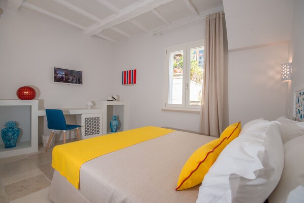 Une chambre d’hôtel avec un lit, un bureau et une chaise. Il y a une fenêtre avec des rideaux, un vase et un téléviseur accroché au mur.