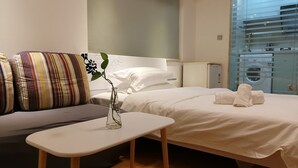 Ein modernes Hotelzimmer mit Bett, einem kleinen Tisch, einer Couch und einem durch eine Glasscheibe abgetrennten Waschbereich.