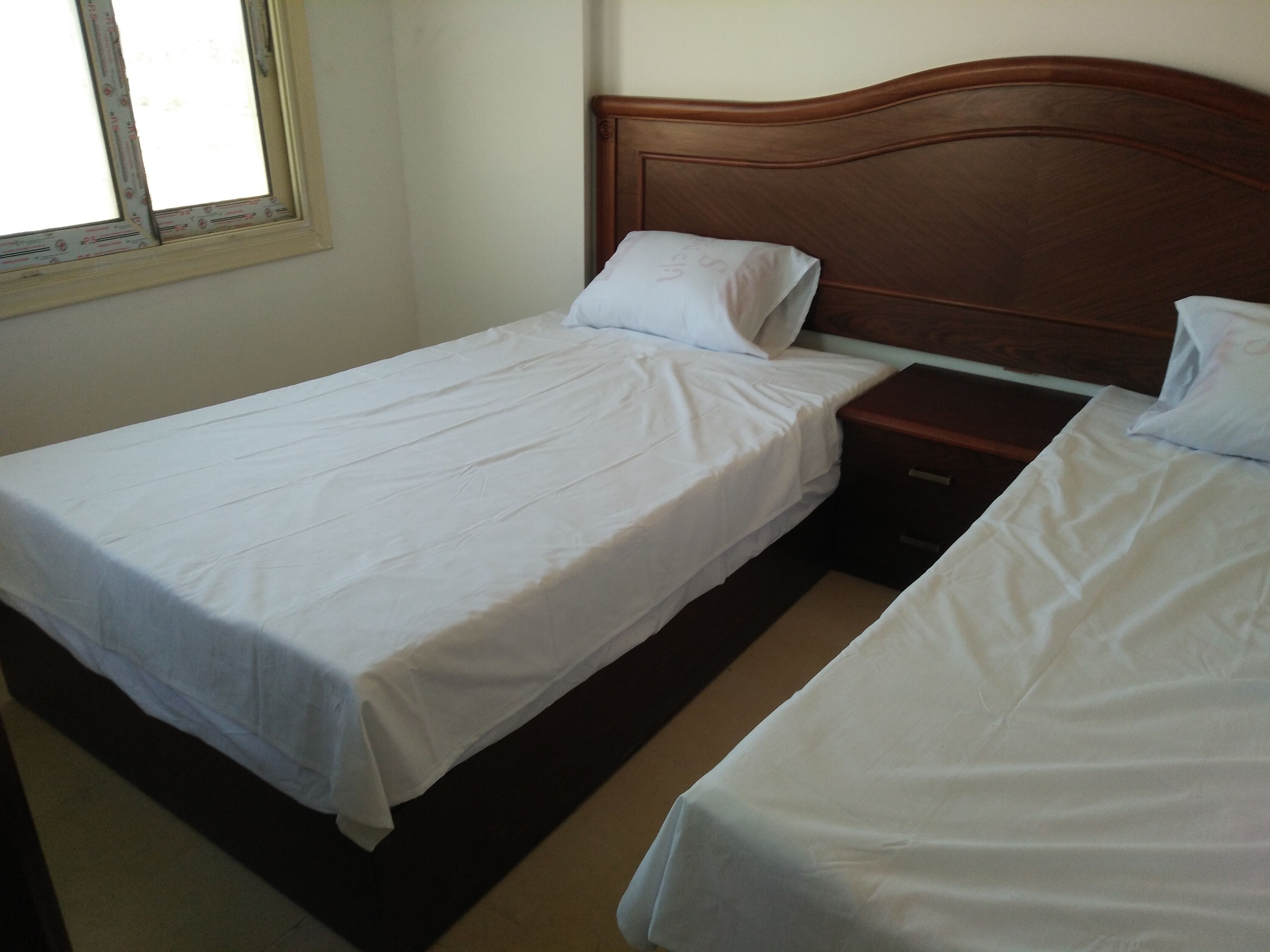 A bedroom with two single beds, a wooden headboard, a nightstand, and a window with blinds.