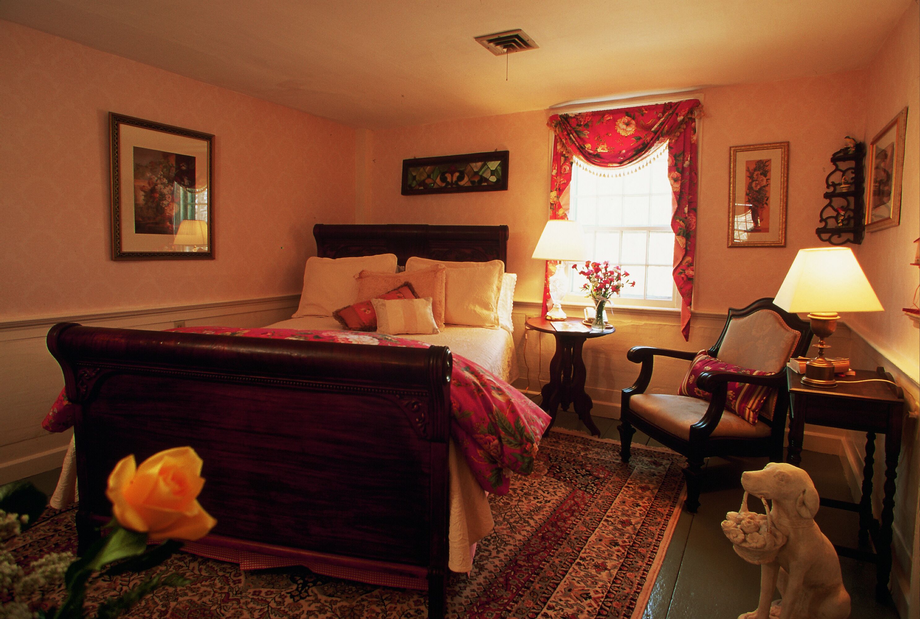 A bedroom with a bed, a chair, a lamp, a vase with flowers, and a dog statue.