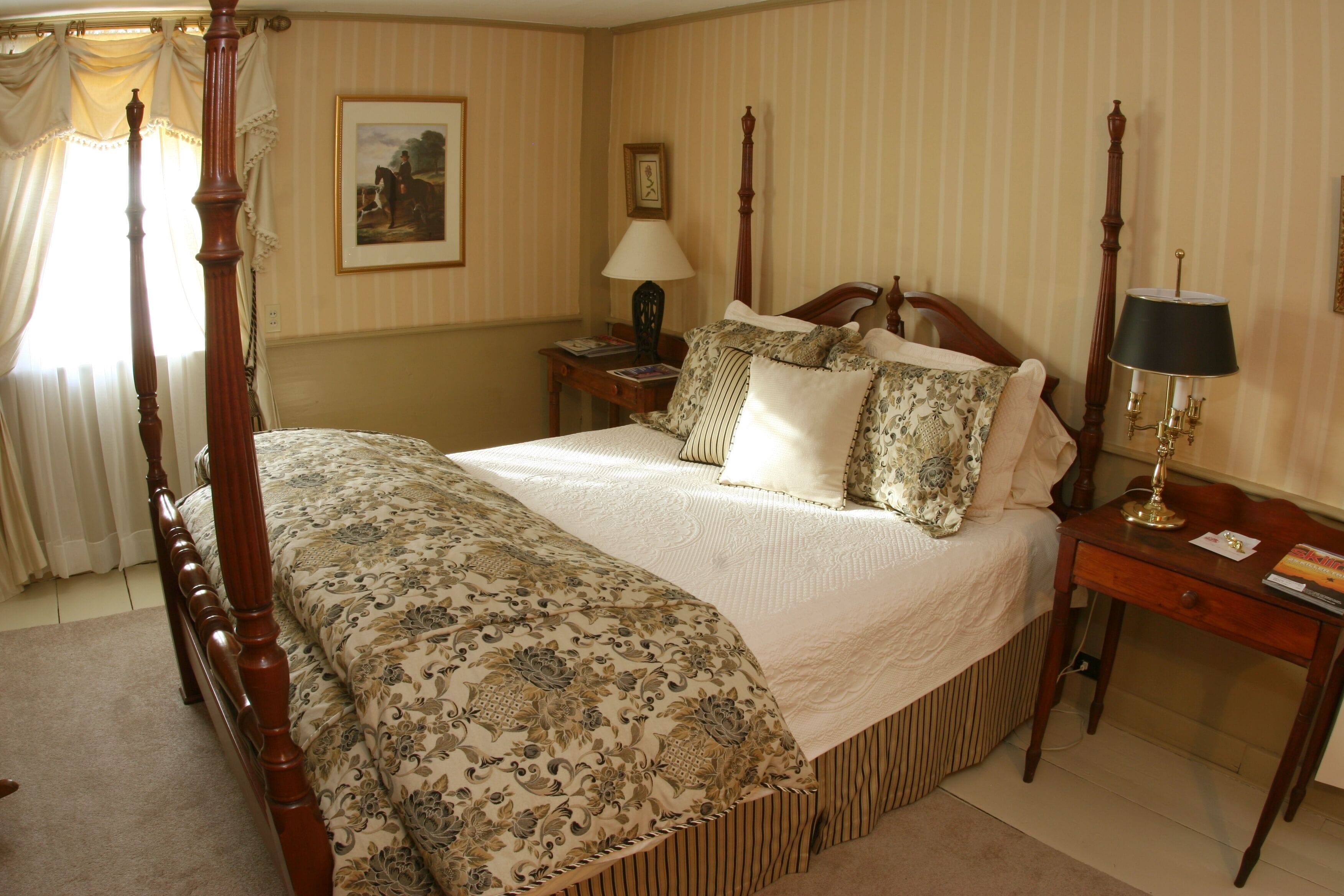 Una cama con dosel con un edredón estampado con motivos florales, una mesita de noche con una lámpara y un cuadro en la pared.