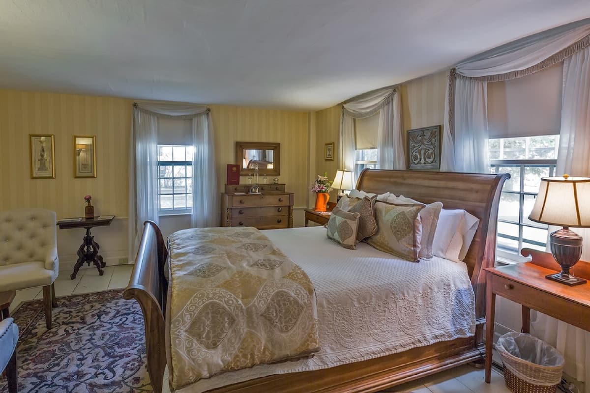 A spacious bedroom with a large bed, a wooden nightstand, a chair, a dresser, and a window with curtains.