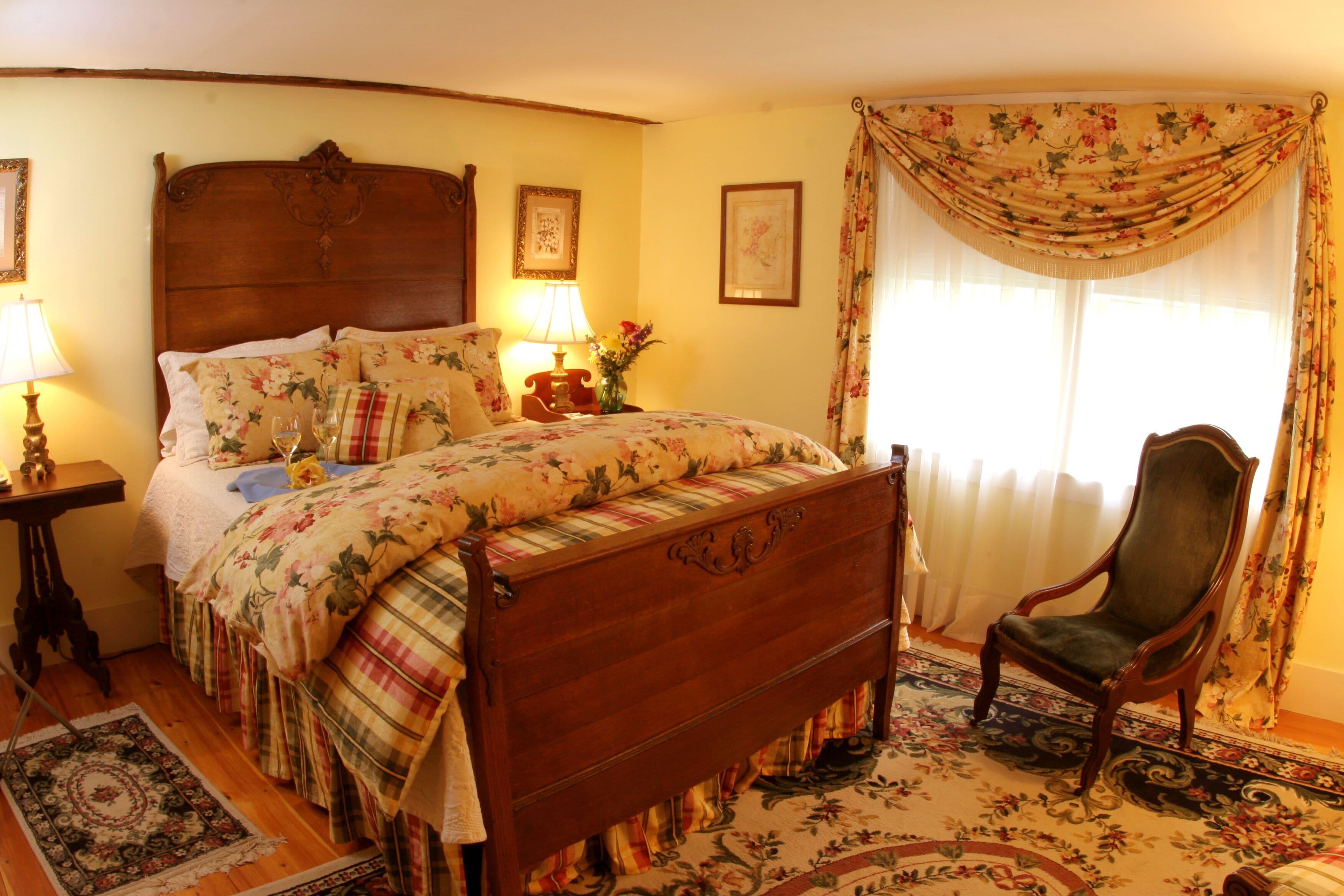 A traditional bedroom with a wooden bed, floral bedding, a chair, and a nightstand with a lamp.