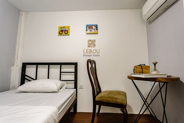 A small, neatly arranged room with a single bed, a chair, a small table with a book and a vase, and two wall-mounted pictures.