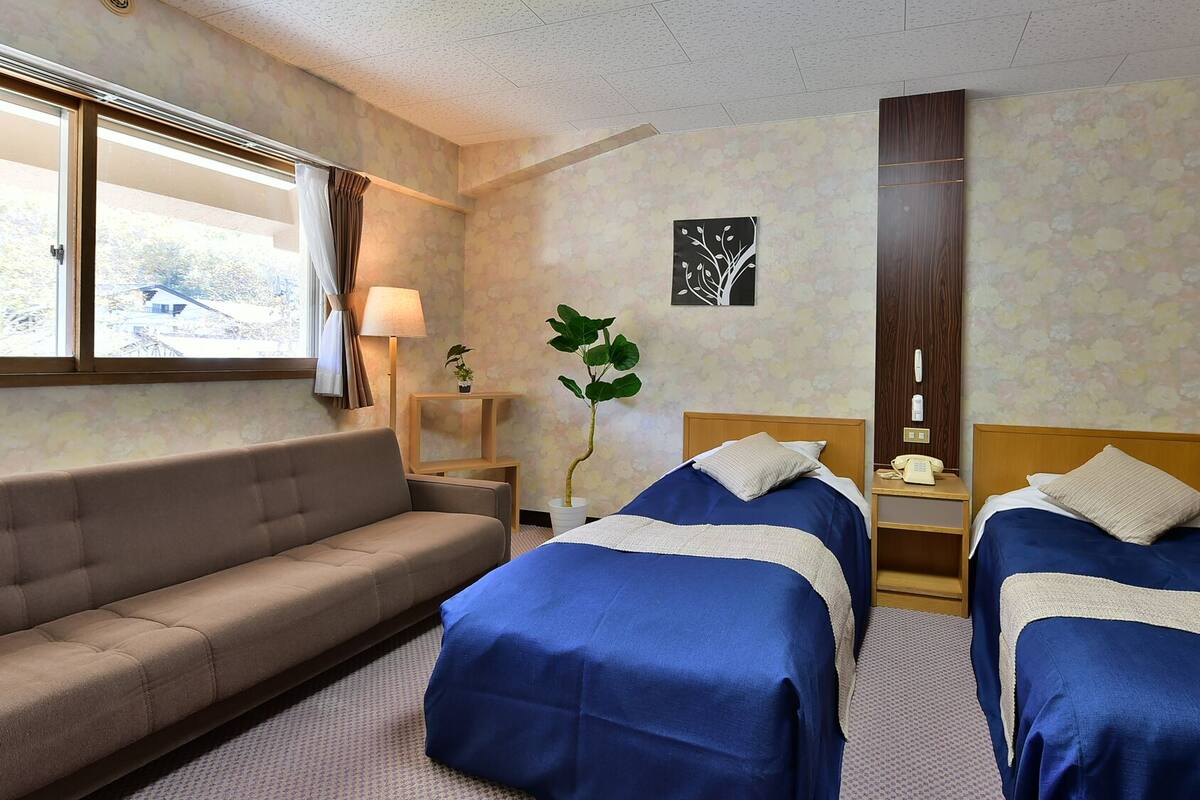 A hotel room with two beds, a sofa, a desk with a lamp, a telephone, and a window with curtains.