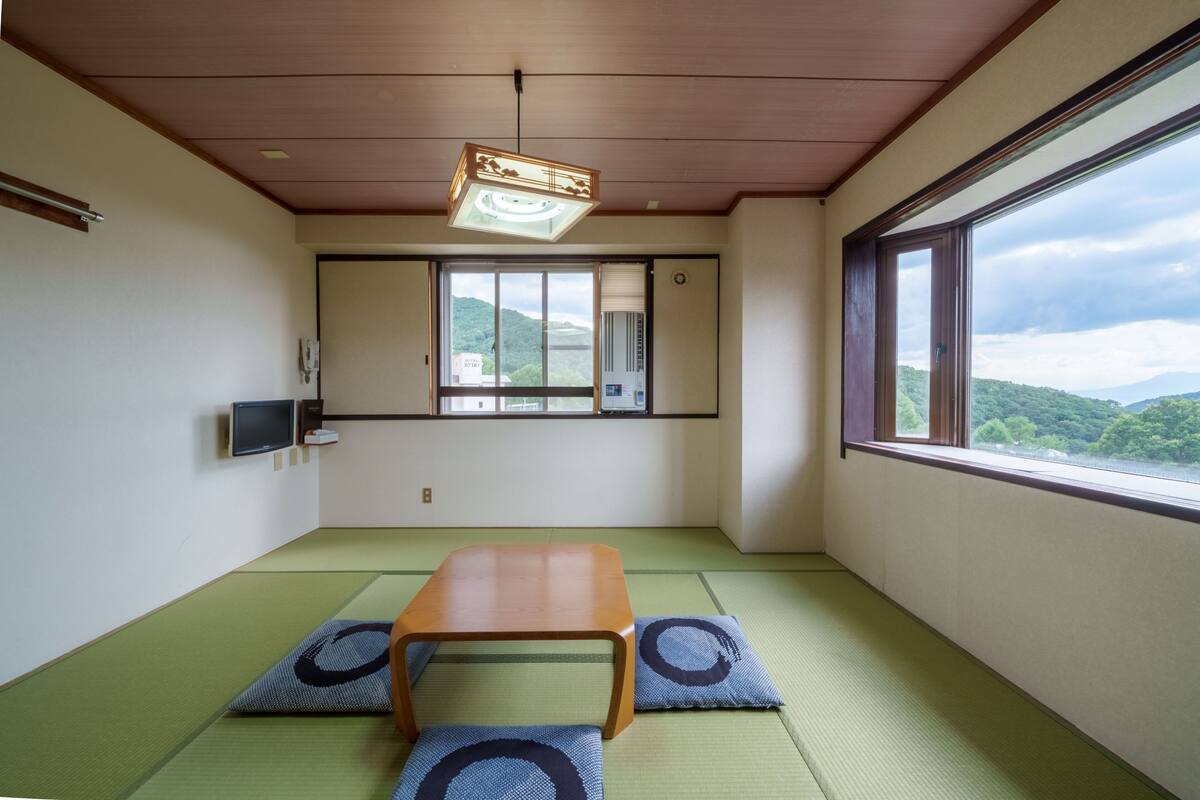 Ein Zimmer mit Tatami-Bodenbelag, einem Holztisch, zwei Tatami-Matten mit einem kreisförmigen Muster, einer Deckenleuchte, einem Fenster mit Blick auf grüne Hügel und einem an der Wand befestigten Fernseher.