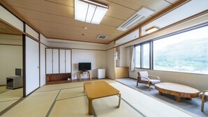 A traditional Japanese-style room with tatami flooring, a television, a small refrigerator, and a view of a mountainous landscape through large windows. - Hotel Tagawa (Yamanouchi)