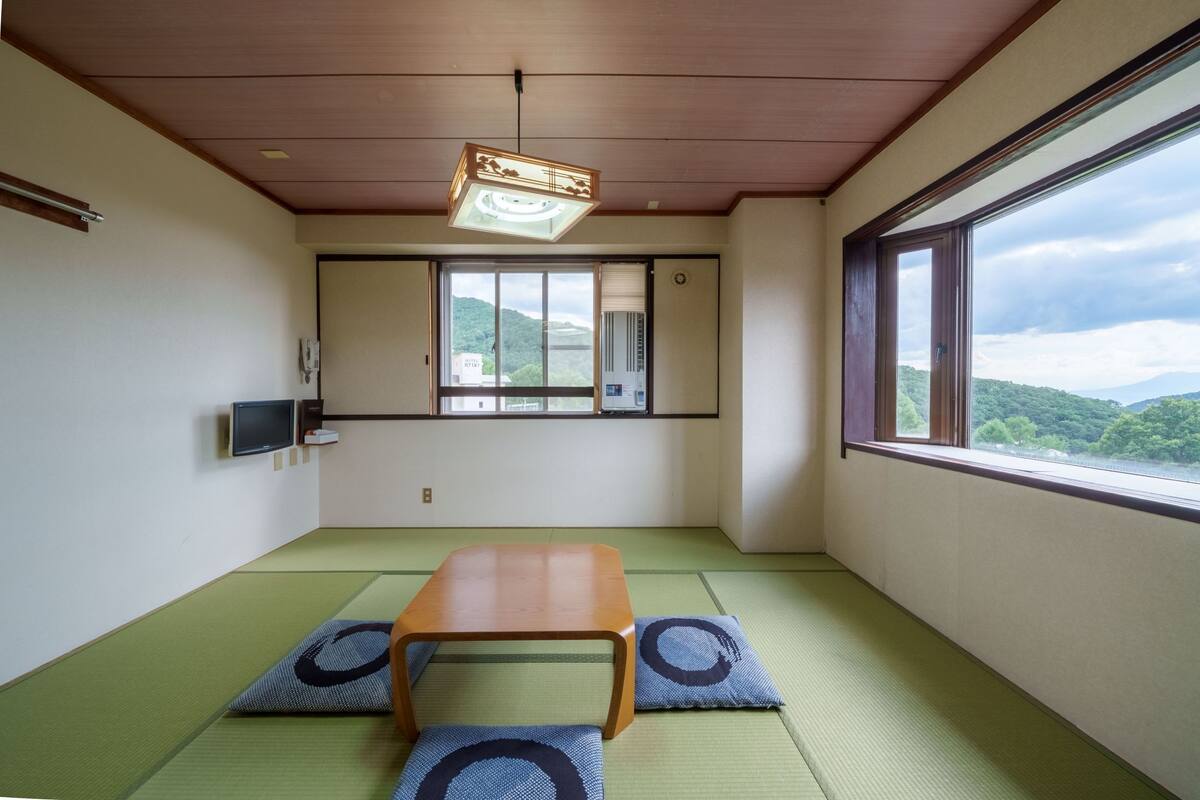 Ein Zimmer mit Tatami-Böden, einem Holztisch, einem Fernseher und einem Fenster mit Blick auf Hügel.