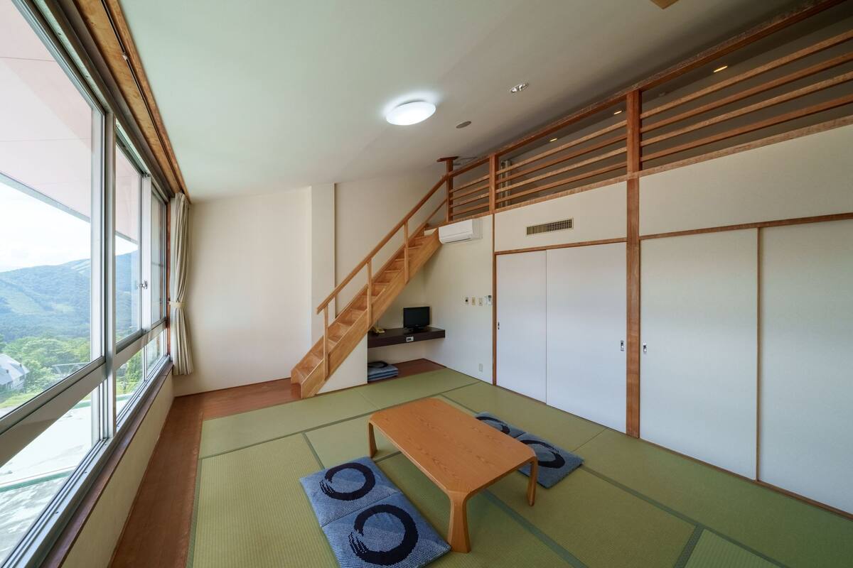 Ein Zimmer mit Tatami-Böden, einem Holztisch, einer Couch und einer Treppe, mit Blick ins Freie.
