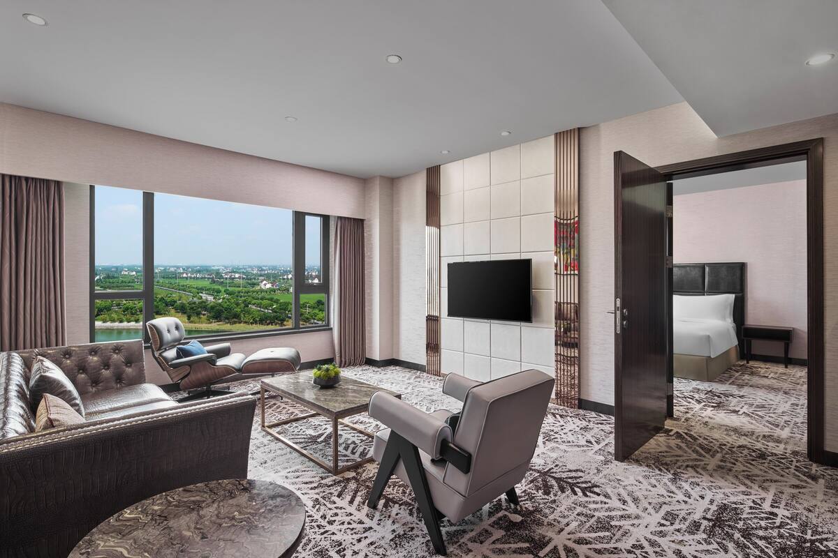 A modern hotel room with a large window offering a city view, a flat-screen TV mounted on the wall, and a comfortable seating area with a sofa and armchairs.
