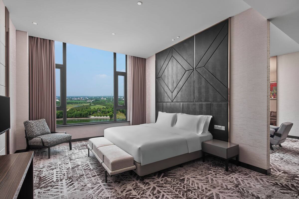 A modern hotel room with a large bed, a seating area with a chair, a TV, and a view of a cityscape.