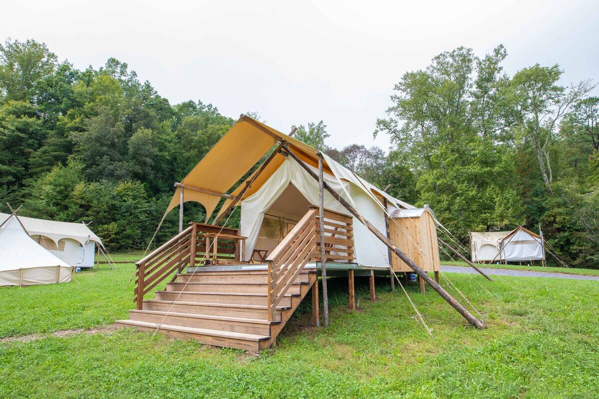 A large, multi-level tent with wooden stairs and a ramp, surrounded by other tents in a grassy area with trees in the background.