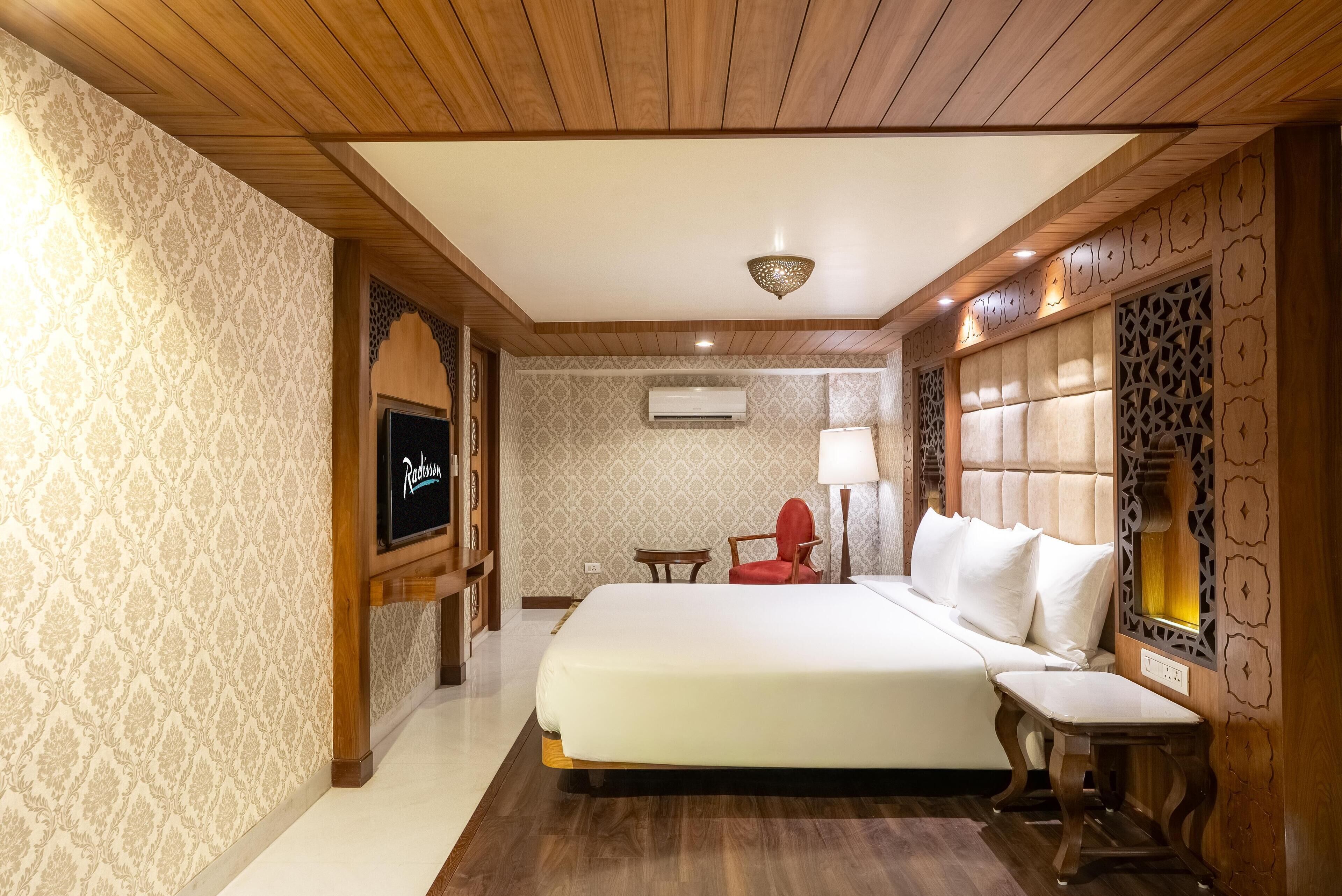 A hotel room with a large bed, a TV, a red chair, and a small table.