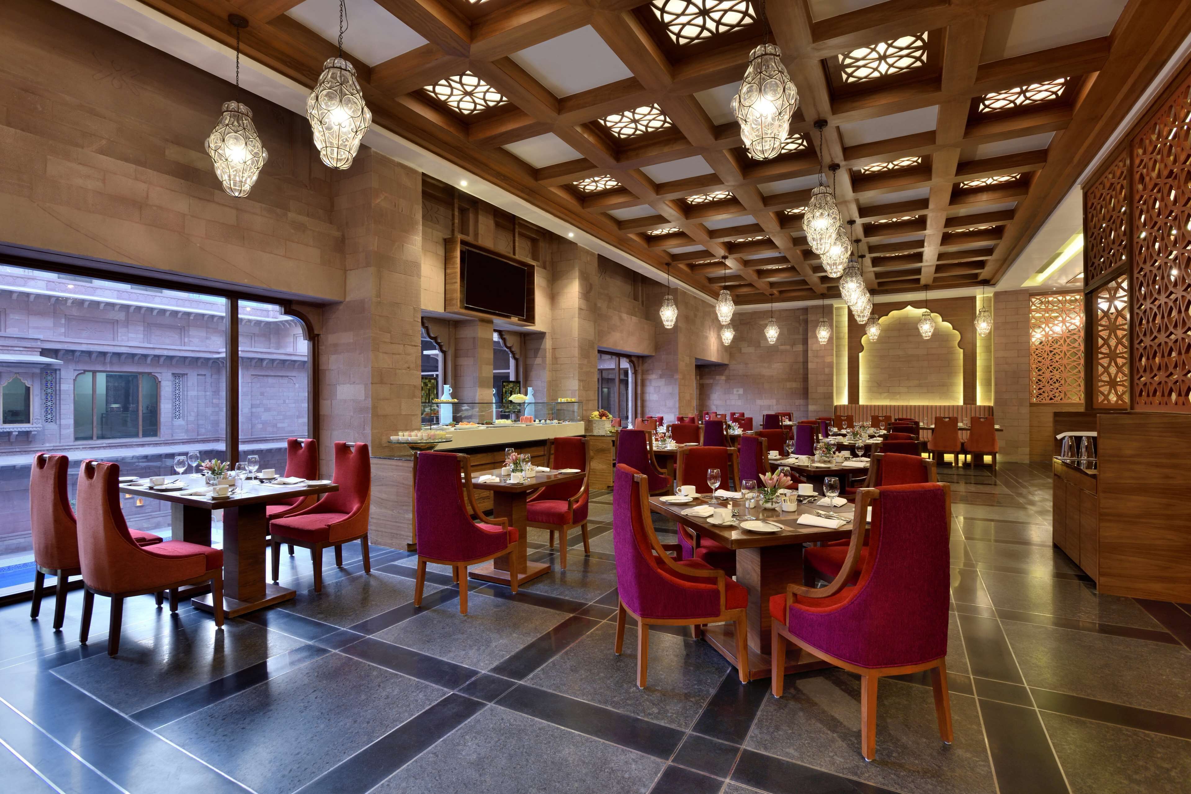 A modern restaurant with wooden ceiling beams, hanging crystal chandeliers, and red upholstered chairs.